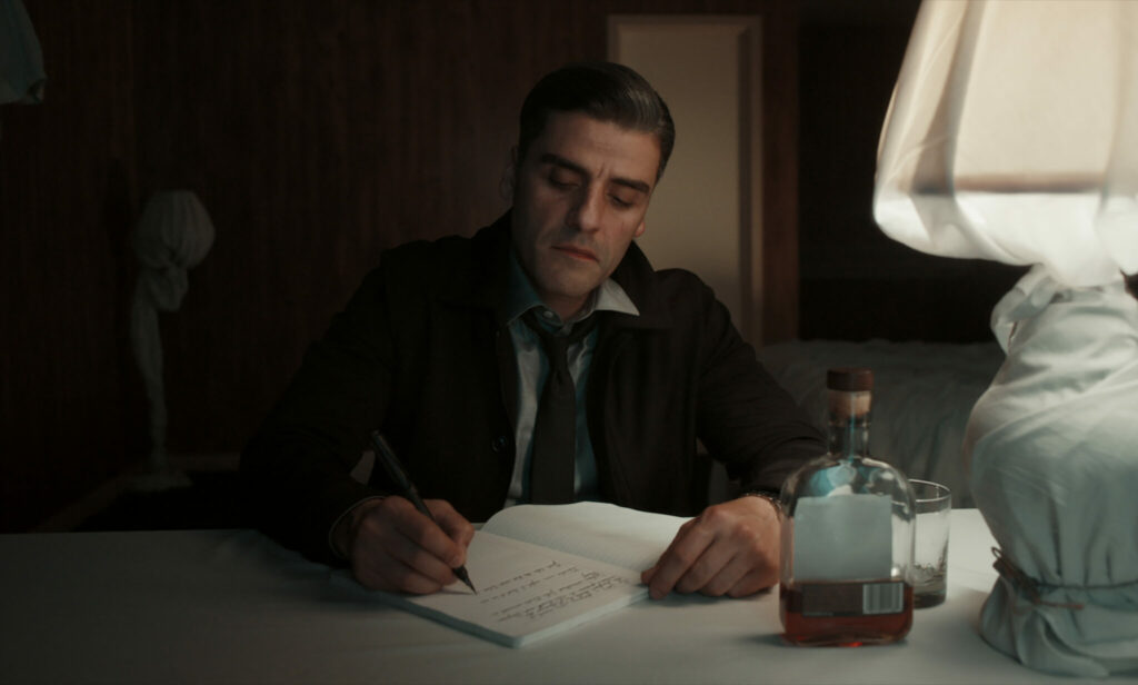 Oscar Isaac sitzt an einem Tisch und führt Tagebuch. Neben ihm steht eine Flasche Whiskey. In The Card Counter