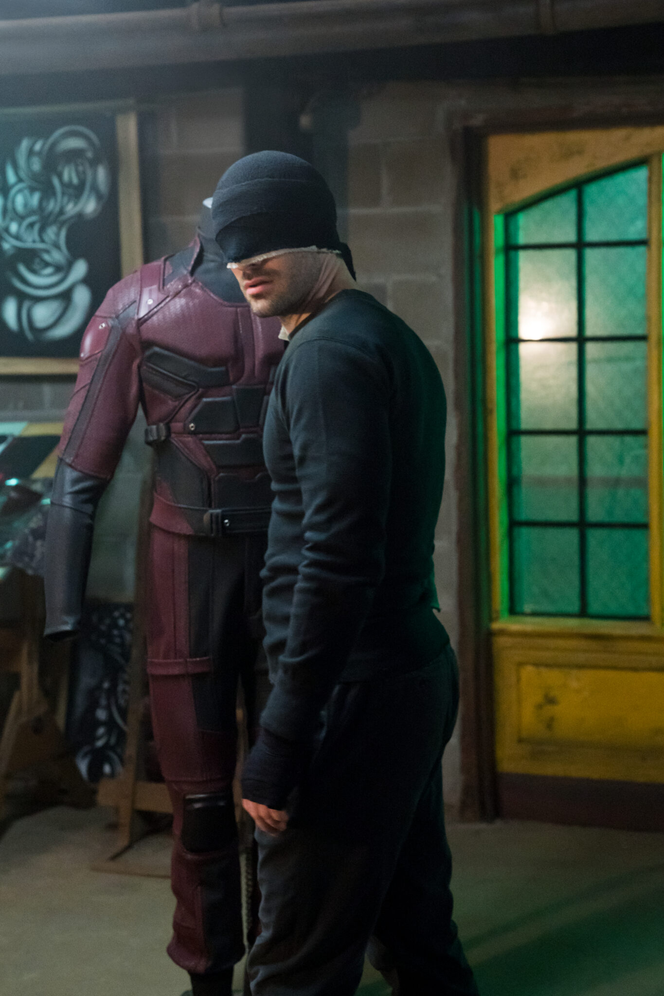 Matt Murdock mit schwarzem Outfit und Tuch über den Augen steht vor einem Ständer auf dem das dunkelrote Daredevil-Kostüm hängt.
