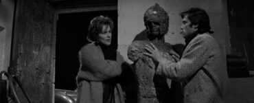 Künstlerin Vivica Lindfors lehnt locker an die Mauer und redet auf Rocker Oliver Reed ein, der Hand an ihre unfertige Skulptur legt - Hammer Horrorfilme.