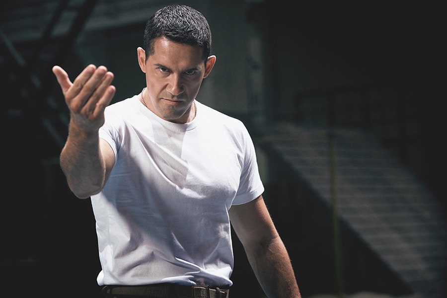 Scott Adkins als Marineoffizier Barton steht in Ip Man 4: The Finale angespannt und mit grimmigem Blick da. Er trägt ein reines weißes T-Shirt und gibt mit seiner Handbewegung zu erkennen, dass er bereit für den Kampf ist.