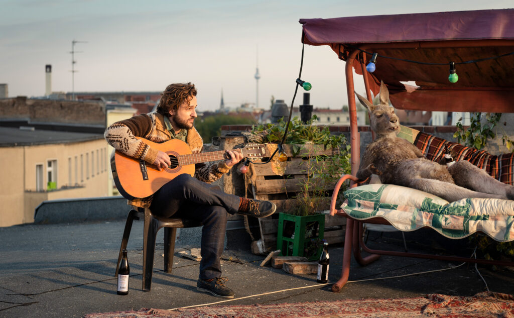 Marc-Uwe und das Känguru sitzen in der Sonne auf einer Dachterasse. Marc spielt die Gitarre während das Känguru entspannt in einer Strandschaukel liegt in Die Känguru-Chroniken