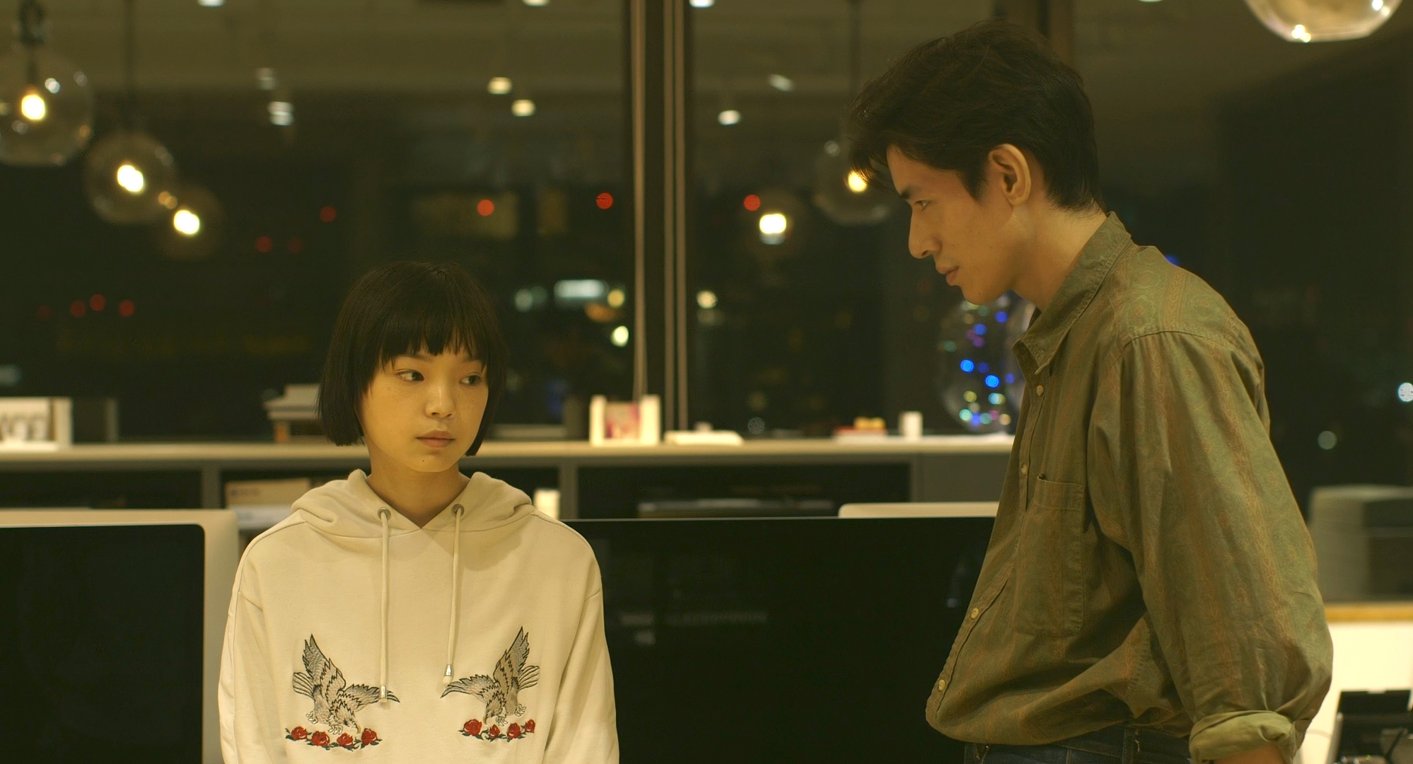 Meiko und Kazuaki nachts in einem Großraumbüro. Sie sitzt mit traurigem Ausdruck und trägt einen weißen Hoodie mit Vogelaufdruck. Er steht rechts neben ihr und trägt ein khakifarbenes Hemd. Im Hintergrund sieht man, dass es außen bereits dunkel ist.