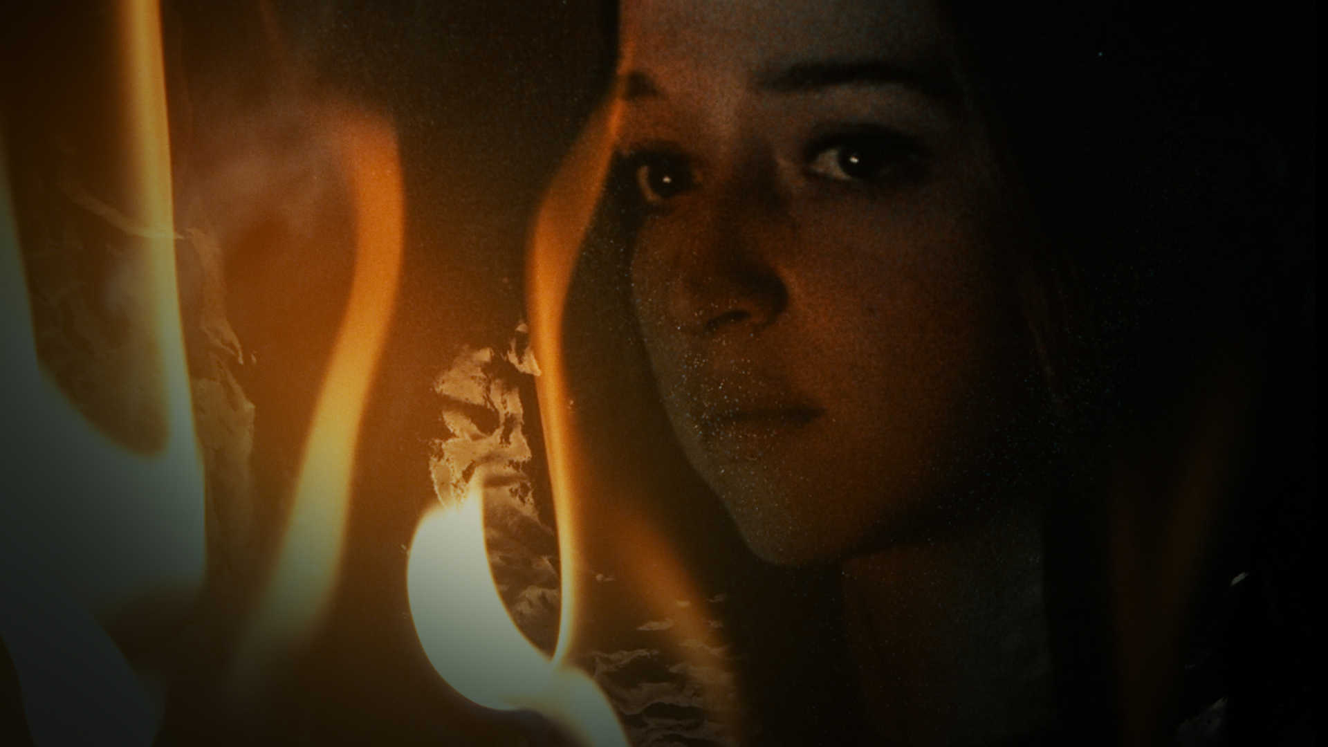 Eine junge Frau mit nachdenklichem Ausdruck im Gesicht in der Nahaufnahme. Links im Bild züngeln vereinzelte Flammen.