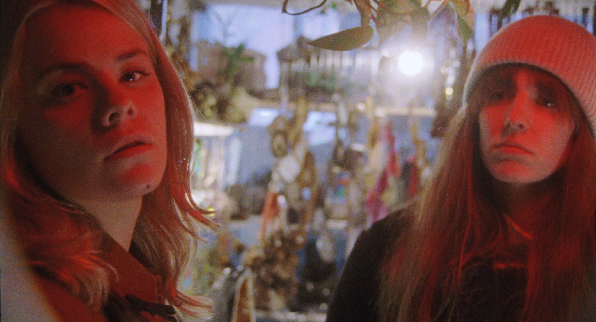 Dasha Nekrasova und Madeline Quinn stehen roterleuchtet in einem Raum. Im verschwommenen Hintergrund leuchtet eine Glühbirne und man sieht einige goldfarbene Gegenstände.