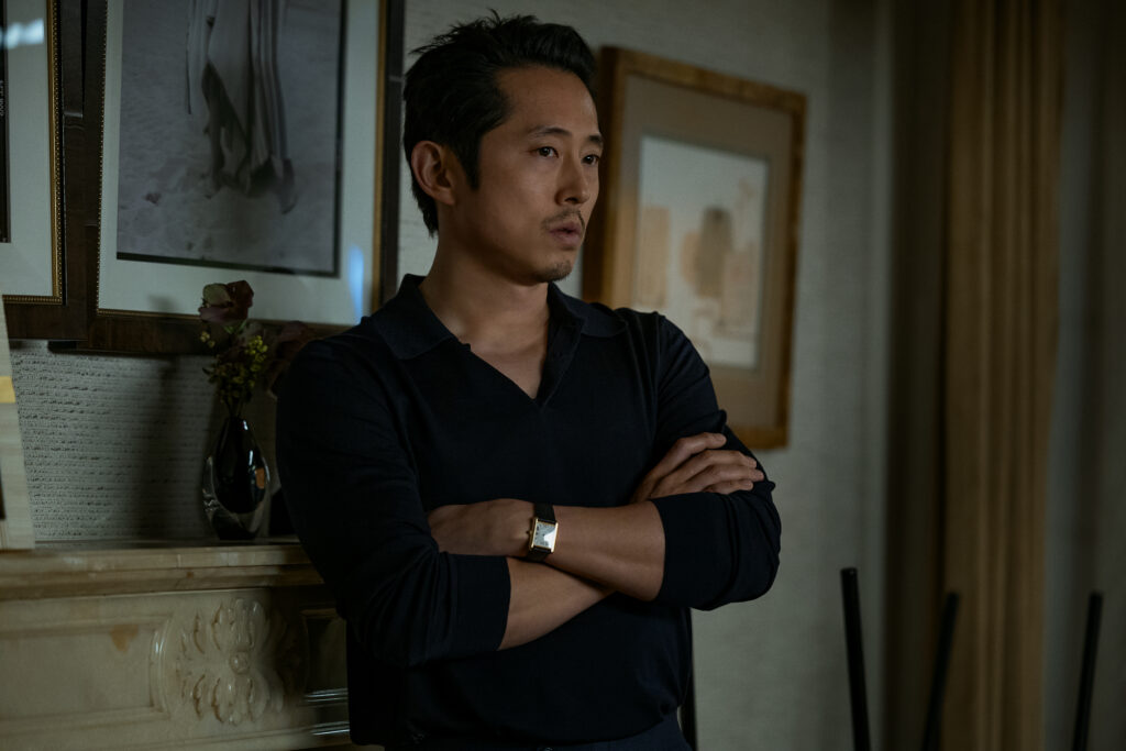 "Man in dark clothing stands indoors with arms crossed, leaning against a mantel with framed pictures and a vase, in a softly lit room with neutral decor and curtains in the background."