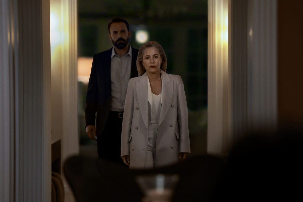 "A woman in a light suit and a man in a dark suit stand in a dimly lit hallway, framed by white columns, facing the camera with serious expressions, suggesting a tense or formal setting."