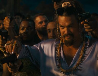 Ein Mann mit lilanem Satin-Hemd und Zöpfen sowie Bart steht vor einer Menschenmenge und hält zwei Waffen in seinen Händen hoch. Es der Schauspieler Jason Momoa als Dante Reyes in Fast & Furious 10.