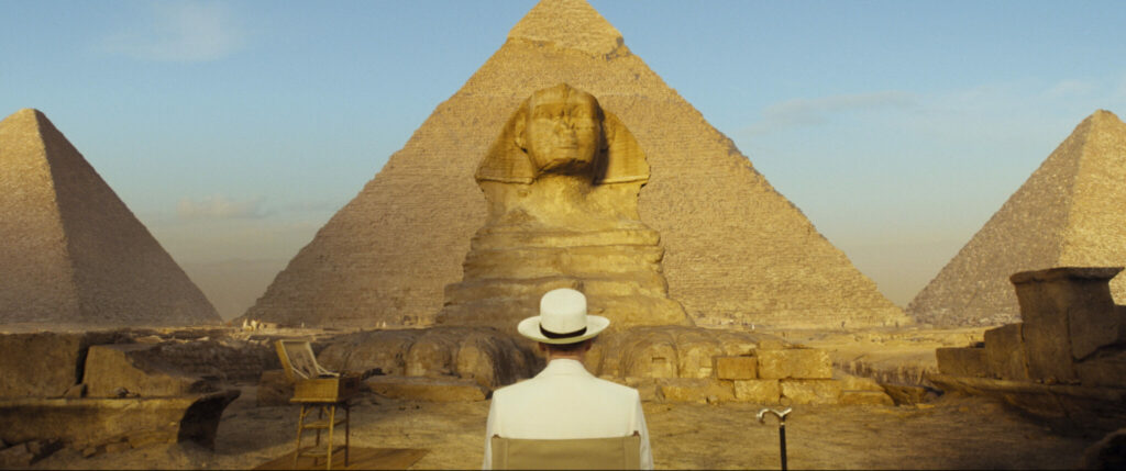 Hercule Poirot (Kenneth Branagh) sitzt abgewandt auf einem Stuhl. Er sieht in die Ferne und betrachtet die Pyramiden, die den Großteil des Bilds ausmachen -Tod auf dem Nil.