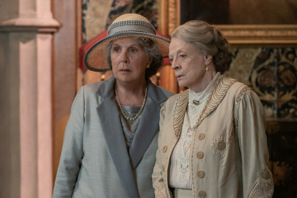Links Penelope Wilton, die einen hellblauen Blazer und einen Hut trägt. Rechts neben ihr Maggie Smith mit einem beigen Jacket, darunter eine weiße Bluse. Eine Szene aus dem Film Downton Abbey 2: Eine neue Ära
