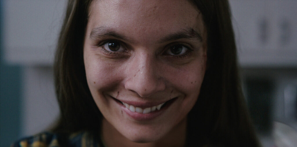 Eine Frau mit brünetten Haaren lacht in die Kamera. Das Lächeln ist allerdings trügerisch, diabolisch und stammt aus dem Film Smile - Siehst du es auch?