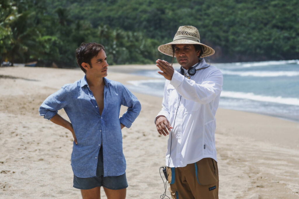 M. Night Shyamalan, der einen Strohhut trägt, gibt Gael García Bernal am Strand Anweisungen für die nächste Szene, gestikuliert mit seinem linken Arm - Old