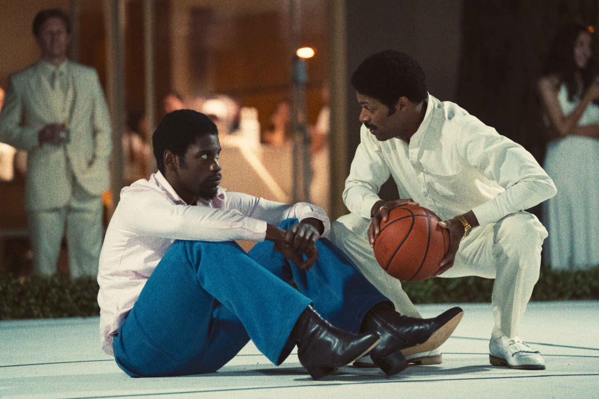 Quincy Isaiah als Basketballlegende Magic Johnson mit seinem Konkurrenten Norm Nixon (DeVaughn Nixon) im Gespräch. Isaiah sitzt auf dem Boden mit blauer Hose und weißem Hemd und Nixon kniet komplett in weiß gewandet vor ihm mit einem Ball in den Händen. Winning Time