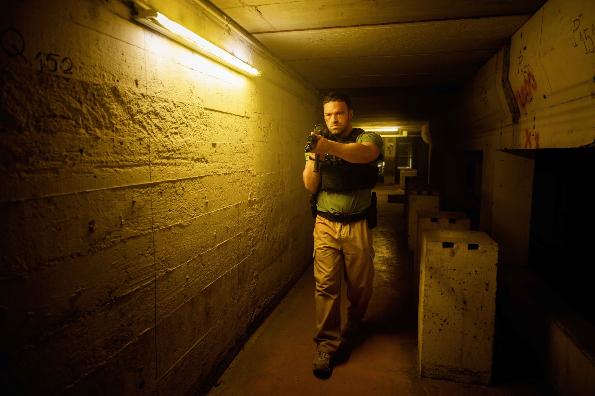 Ein Polizist mit Schussweste geht mit erhobener Waffe durch einen engen, dunklen Kellergang. AKA