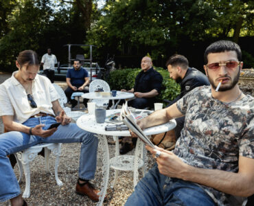 Einige Gangster sitzen an einem kleinen, weißen Tisch mit Handys oder Zeitschriften in den Händen, manche rauchen zudem.