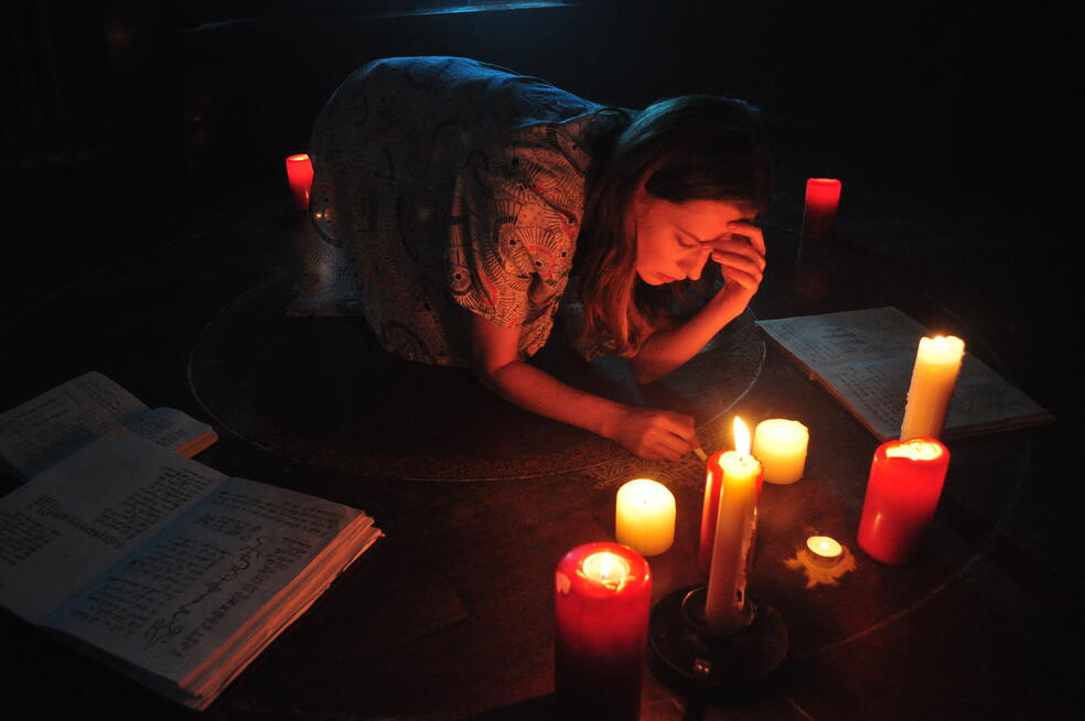 Sophia zeichnet etwas auf den Boden, um sie herum sind brennende Kerzen und aufgeschlagene Bücher verteilt | A Dark Song