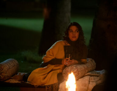 Eine der Freundinnen aus Hush Hush liegt zwischen Bäumen auf einer Sänfte im Dunkeln, vor ihr ein kleines Lagerfeuer - Neu bei Prime im September 2023