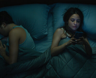 Ein Mann und eine Frau liegen im Bett, während der Mann zu schlafen versucht, ist die Frau am Handy - Neu bei Prime im Februar 2023