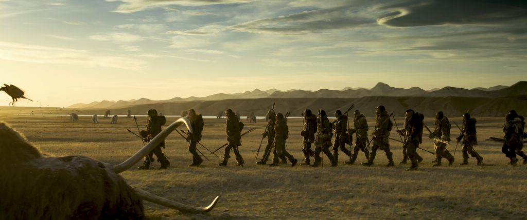 Die Männer des Stammes ziehen mit Speeren bewaffnet zur Jagd, im Vordergrund ein Teil eines erlegten Mammuts