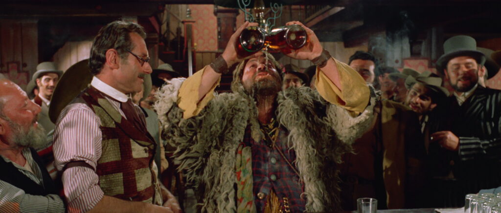 Whiskey, gespielt von Roberto Carmadiel, trinkt an der Theke des Saloons von Blackstone Hill gleich aus zwei Flaschen auf einmal. Saloonbesitzer Pedro, gespielt von Andrea Bosic, steht staunend daneben.