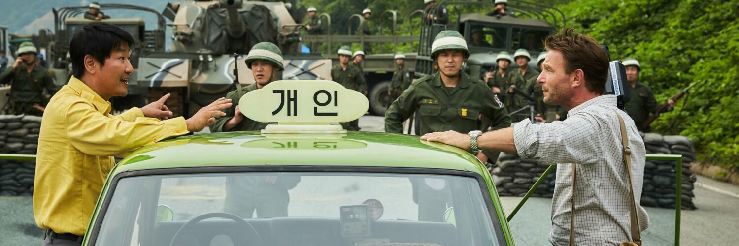 Der Taxifahrer steht auf der linken Seite seinem Fahrgast Jürgen Hinzpeter gegenüber. Zwischen ihnen steht das grüne Taxi und im Bildhintergrund das Militär.