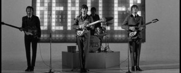 Auf einem Schwarz-weiß-Bild sieht man links Paul McCarney, in der Bildmitte George Harrison, hinter ihm Ringo Starr am Schlagzeug und rechts John Lennon. Hinter dem Schlagzeug leuchtet ein "The Beatles"-Schriftzug.