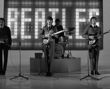 Auf einem Schwarz-weiß-Bild sieht man links Paul McCarney, in der Bildmitte George Harrison, hinter ihm Ringo Starr am Schlagzeug und rechts John Lennon. Hinter dem Schlagzeug leuchtet ein "The Beatles"-Schriftzug.