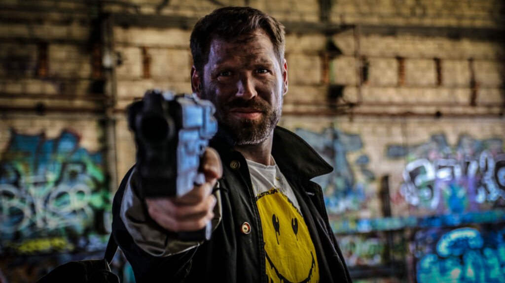 Der Protagonist Alex blickt in RIchtung der Kamera und hält die Mündung seiner Waffe nach vorne gerichtet. Er ist in einer Nahaufnahme positioniert, trägt eine Jacke und ein weißes T-Shirt mit einem gelben Smiley. Im Hintergrund ist Mauerwerk samt Graffiti zu sehen.