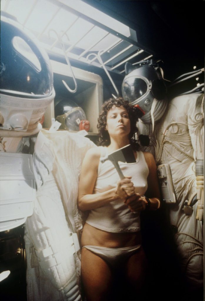 Sigourney Weaver steht als Ellen Ripley leicht bekleidet mit einer Axt in der Hand zwischen an Haken hängenden Astronautenanzügen in einer Ecke in Alien – Das unheimliche Wesen aus einer fremden Welt