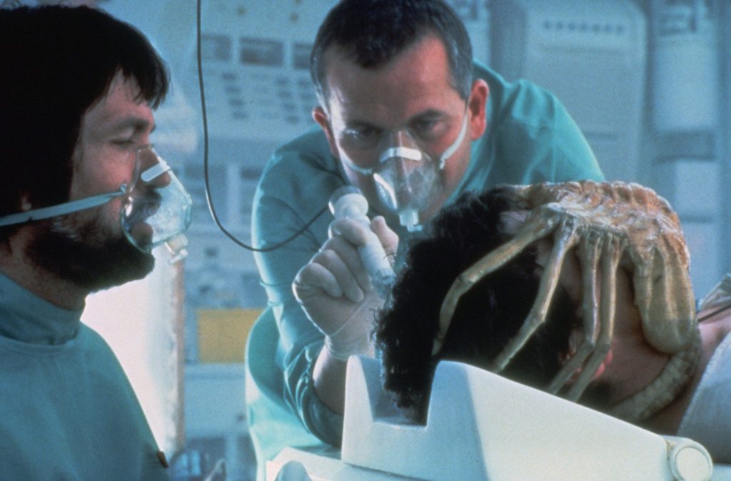 Tom Skerritt und Ian Holm untersuchen mit Atemmasken und Kitteln den vor ihnen liegenden John Hurt, dessen Gesicht vom Facehugger umschlungen wird | Alien – Das unheimliche Wesen aus einer fremden Welt
