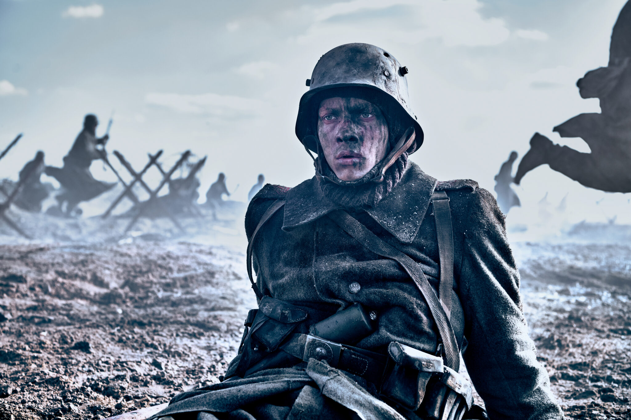 Felix Kammerer als Soldat Paul Bäumer in Im Westen nichts Neues. Er sitzt mit fassungslosem Ausdruck auf dem Schlachtfeld. Hinter ihm sieht man weitere Soldaten zwischen Barrikaden.