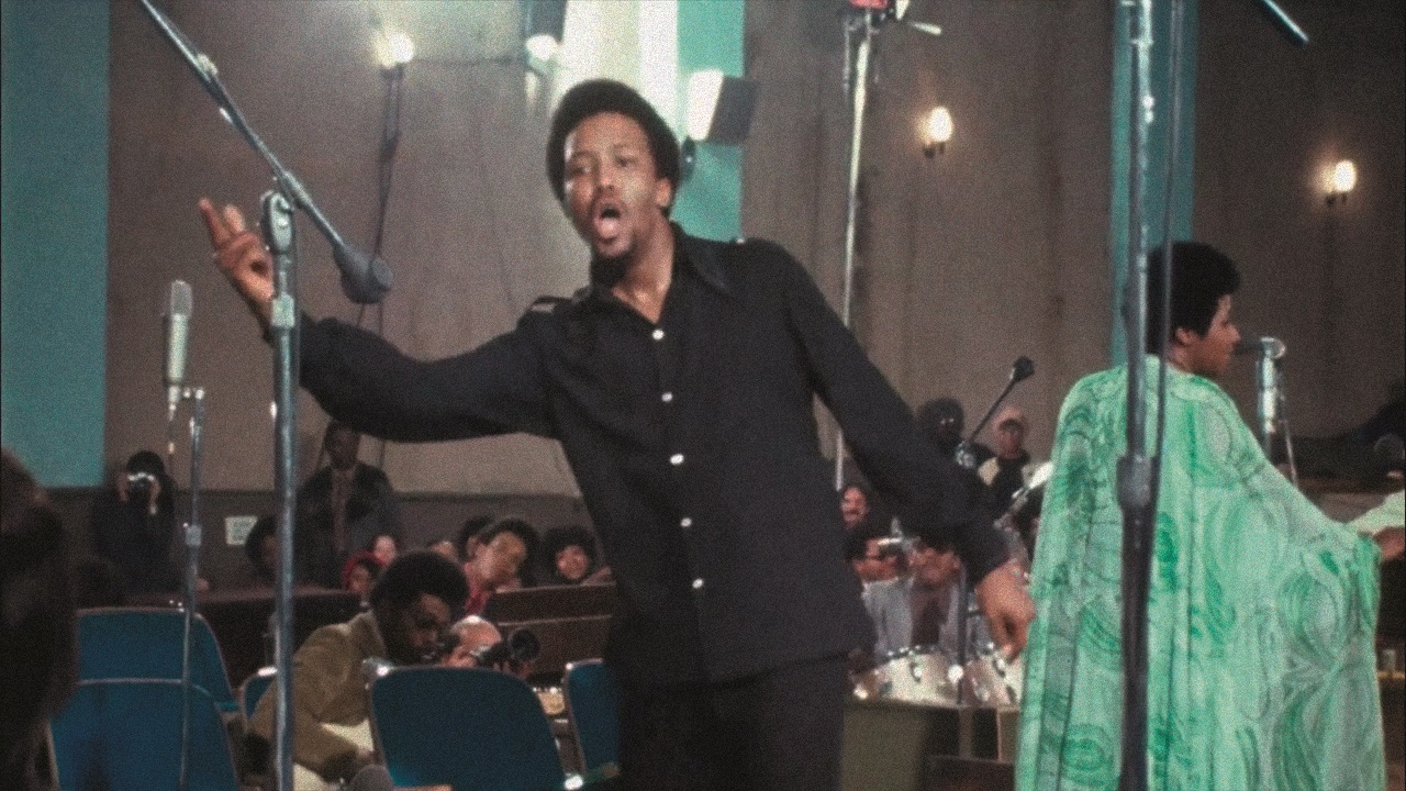 Im Vordergrund ist der Chorleiter Alexander Hamilton zu sehen, wie er seinen Chor animiert. Im Hintergrund steht Aretha Franklin.