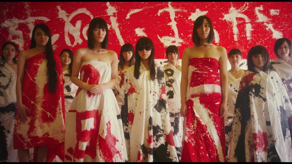 Vor einer rot angestrichenen und weiß beschrifteten Wand blicken Frauen und farblich reduzierten Kleidern in Richtung des Publikums. Rote farbe dominiert das Bild. Die weißen Kleider der Damen sind mit Rot oder Schwarz bemustert.