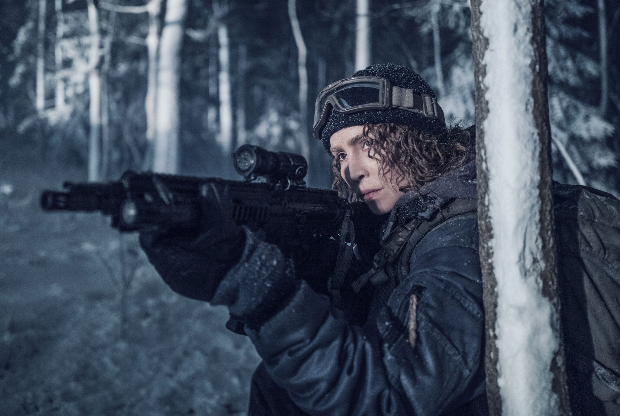 Noomi Rapace in Operation Schwarze Krabbe hinter einem Baum mit Gewehr im Anschlag und im dunklen, wintertauglichen Outfit.