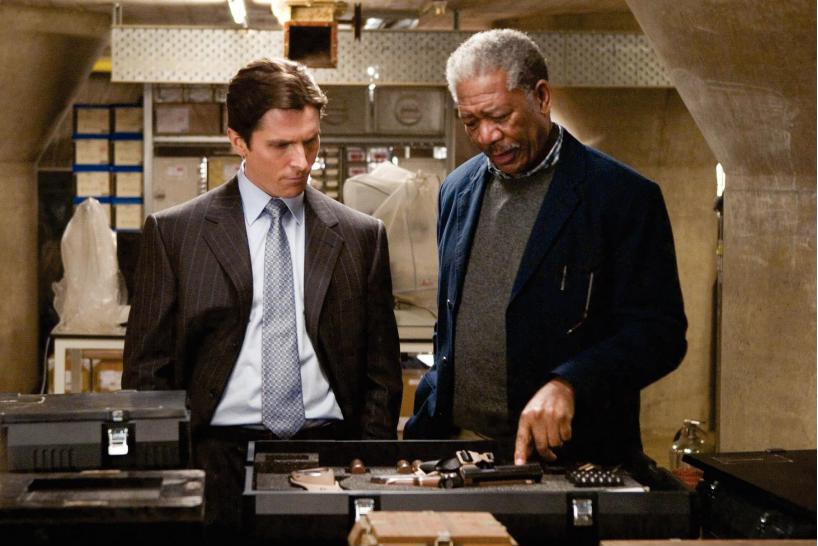 Christian Bale und Morgan Freeman stehen vor einem Tisch. Freeman zeigt auf einige Gadgets in einem Koffer.
