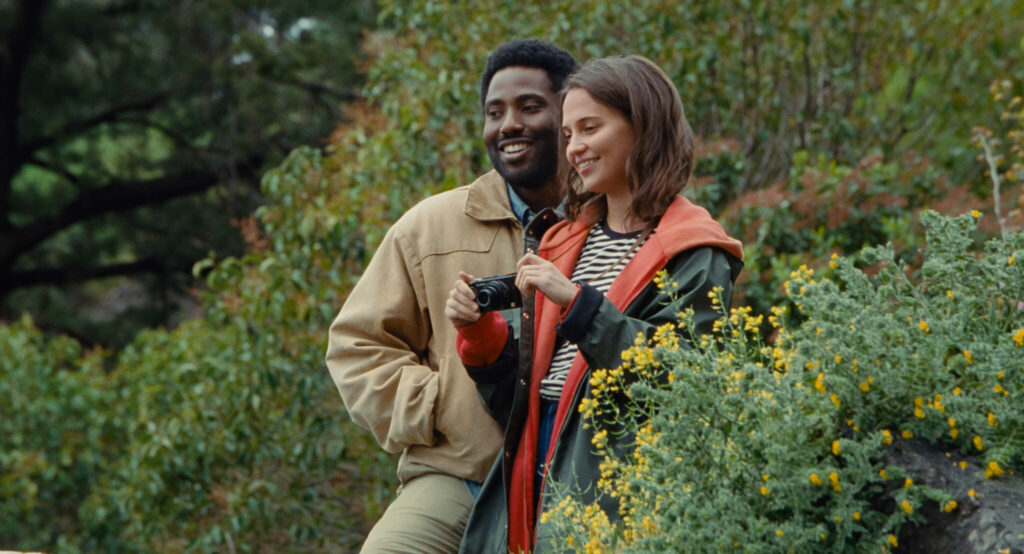 Beckett (John David Washington) und April (Alicia Vikander) stehen in der Natur, während ihres Urlaubs in Griechenland. Es sind einige Pflanzen und Büsche um sie herum zu erkennen. April hält eine Kamera in beiden Händen. Beide lächeln glücklich. 