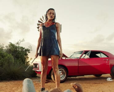 Christina Ochoa als Grace steht in "Blood Drive" blutbespritzt und in einem blauen Kleid über der Leiche eines Mannes. Sie hält einen spitzen Metallstab in der Hand und im Hintergrund ist ihr rotes Auto zu sehen.