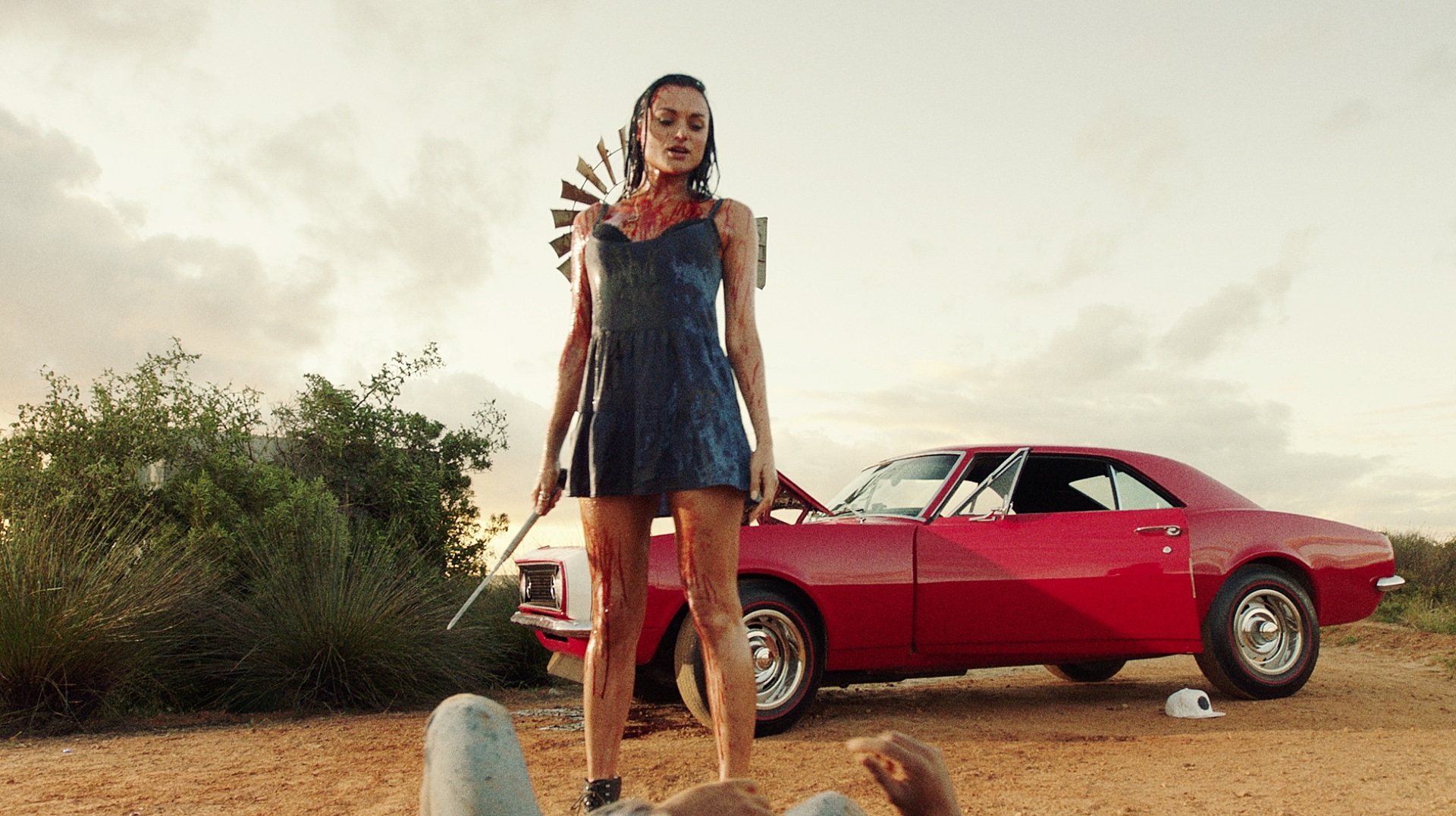 Christina Ochoa als Grace steht in "Blood Drive" blutbespritzt und in einem blauen Kleid über der Leiche eines Mannes. Sie hält einen spitzen Metallstab in der Hand und im Hintergrund ist ihr rotes Auto zu sehen.