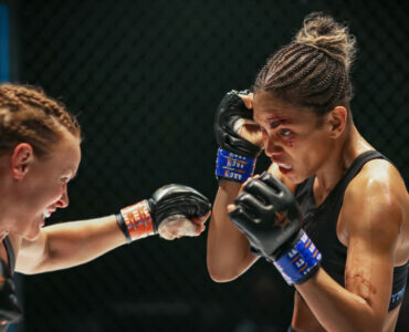 Ein Boxkampf zwischen zwei Sportlerinnen, eine in verteidigender Haltung, die andere während einem Schlag. Bruised