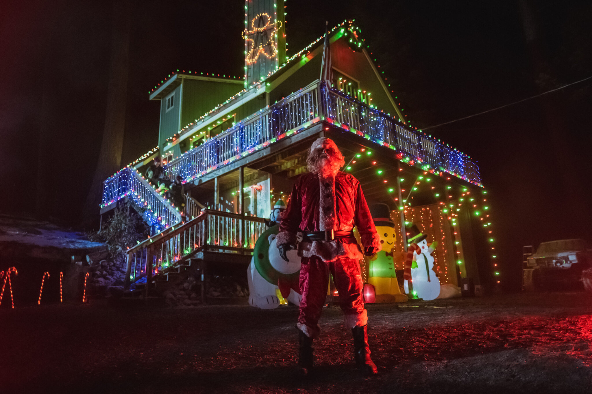 Im Film Christmas Bloody Christmas steht Abraham Benrubi in tiefster Nacht mit dem blutigen Kostüm eines Weihnachtsmannes vor einem grell-bunt weihnachtlich dekoriertem Haus.