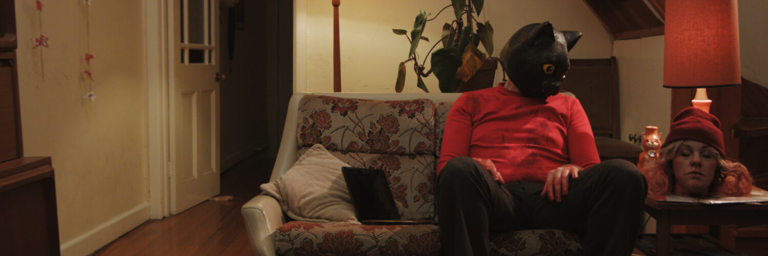 Matthew C. Vaughan sitzt als Ted in Cat Sick Blues mit Katzenmaske auf dem Kopf auf einem Sofa