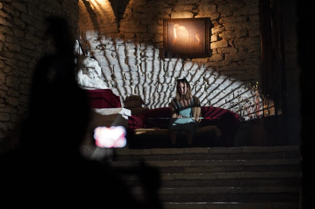 Im gefächerten Licht eines großen Fensters sitzt die blinde Rebecca auf einem Sofa im Flur des Schlosses, zu ihren Füßen eine steinerne Treppe - Castle Freak