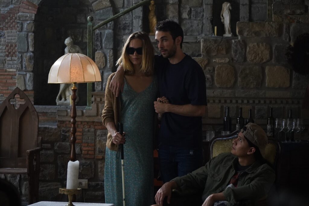 John hält Rebecca, die ihre Brille trägt und ihren Blindenstock in der Hand hält, im Arm, als sie ihre Freunde in einem der Wohnzimmer des Schlosses willkommen heißen - Castle Freak