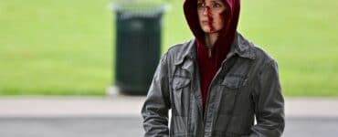 Ava (Jessica Chastain) steht an der Straße und beobachtet etwas. Sie trägt eine dunkelgrüne Jacke und einen bordeauxroten Hoodie. Die Kapuze des Hoodie ist übergezogen, verdeckt allerdings nicht die vielen Wunden in Avas Gesicht.