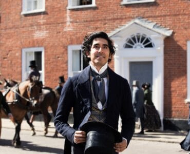 David Copperfield, gespielt von Dev Patel, steht vornehm gekleidet mit dem Zylinder in den Händen auf einer belebten Londoner Straße und blickt lachend um sich.