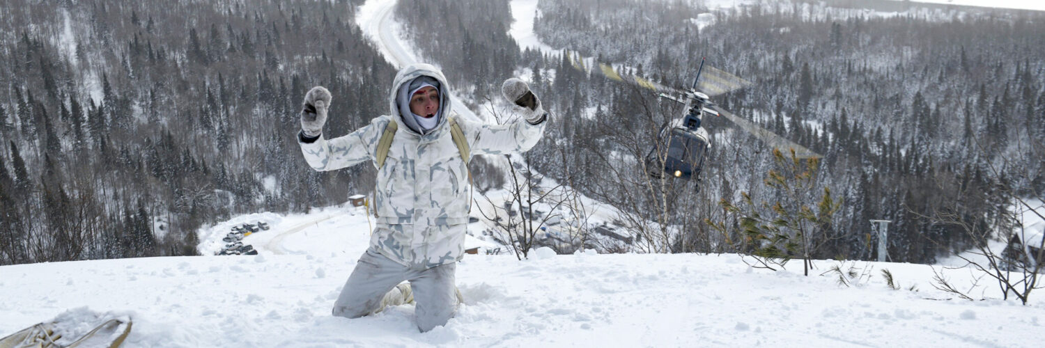 Ein Drogenkurier, ganz in weiß gekleidet, kniet auf einem schneebedeckten Hügel mit erhobenen Händen. Im Hintergrund sieht man die verschneite Wildnis und einen Helikopter herannahen. Crisis.
