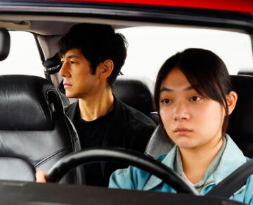 Misaki (Toko Miura) sitzt am Steuer des roten Saabs 900, während Yusuke (Hidetoshi Nishijima) hinter ihr auf der Rückbank sitzt und seitlich aus dem rechten Fenster blickt. Beide wirken angestrengt und erschöpft. Zudem ist die Umgebung außerhalb des Autos nicht erkennbar.