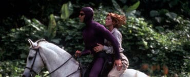 Billy Zane alias Das Phantom reitet auf einem weißen Pferd, hinter ihm sitzt Kristy Swanson in einem beigefarbenen Abenteureroutfit und schulterlangen rotbraunen Haaren. Im Hintergrund ist der Dschungel zu sehen.