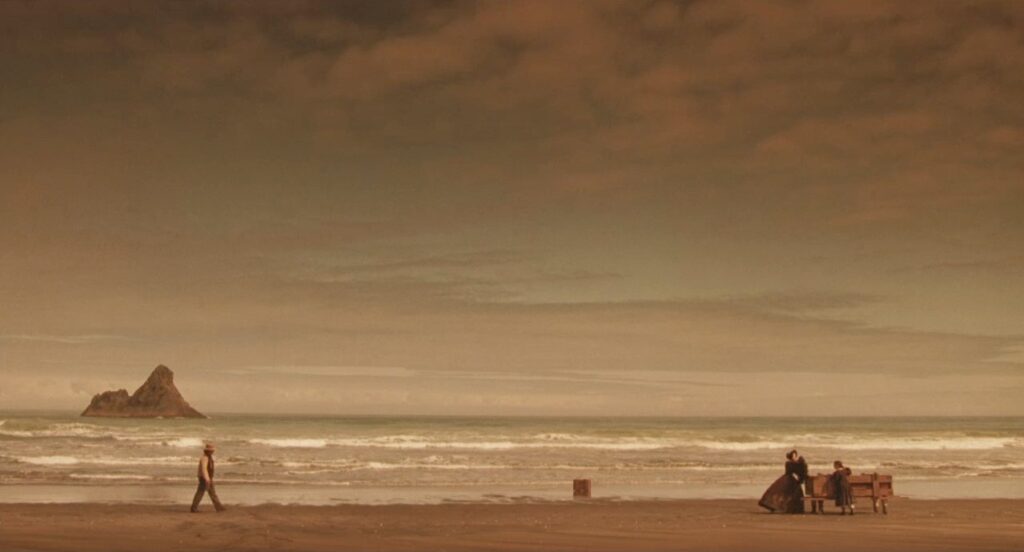 Ein weites Bild von Strand und Himmel. Unten vor dem Meer stehen Holly Hunter und Anna Paquin am Piano ganz am rechten Rand. Etwas weiter links von ihnen steht eine Kiste im Wasser. Links kommt Harvey Keitel auf sie zu geschritten. Hinter ihm liegt im Meer eine kleine Felsinsel.