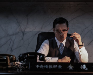 Direktor Kim (Lee Byung-hun) sitzt in Das Attentat - The Man Standing Next nachdenklich und rauchend in seinem Büro, vor ihm stehen zwei Telefonapparate mit Wählscheibe.
