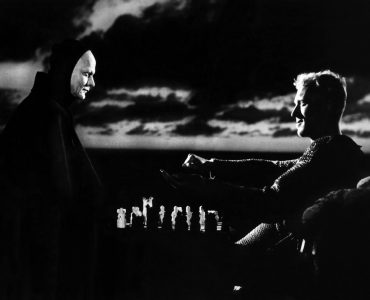 Der Tod (Bengt Ekerot) sitzt links im Bild und spielt Schach mit A dem ihm gegenüber sitzenden Antonius Block (Max von Sydow). Im Hintergrund ballen sich dunkle Wolken am Himmel.